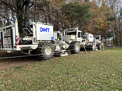 Zwei Vibro-Trucks hintereinander fahren am Waldrand auf einem Waldweg.