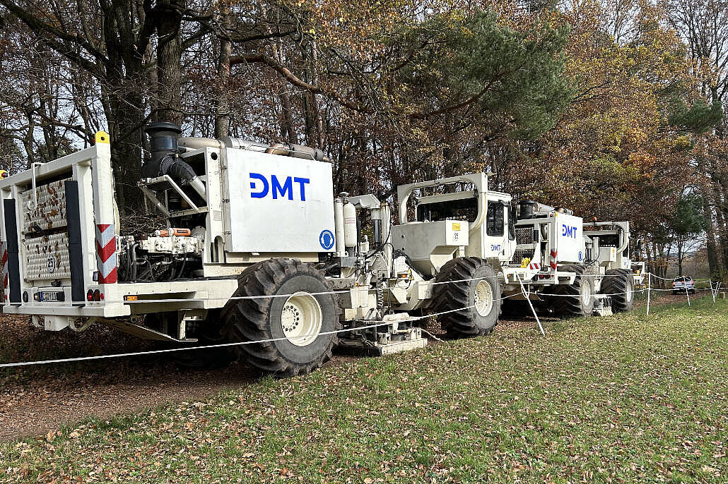 Zwei Vibro-Trucks hintereinander fahren am Waldrand auf einem Waldweg.