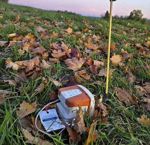 Messgerät auf einer Wiese mit Laub. Direkt daneben steckt eine Markierung in Form eines langen Stabs im Boden.